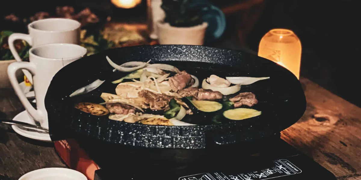 Korean BBQ grilling meat, onions, and zucchini slices on a tabletop pan with dim lighting.