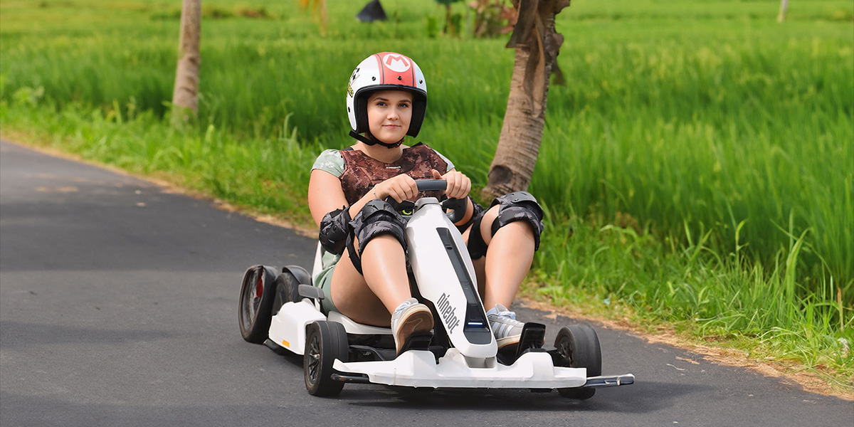 Young woman wearing helmet and knee pads riding a white go-kart on a paved path next to green grass.