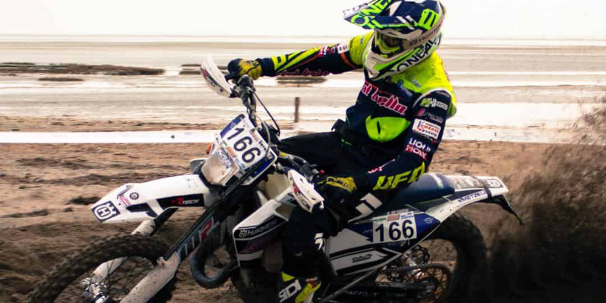 Dirt bike rider wearing yellow and black gear with helmet, racing on sandy terrain kicking up dirt.