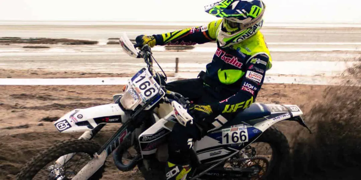 Dirt bike rider wearing yellow and black gear with helmet, racing on sandy terrain kicking up dirt.