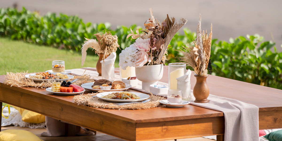 Outdoor wooden table set with breakfast plates, fruit, pastries, candles, and floral centerpieces on placemats.