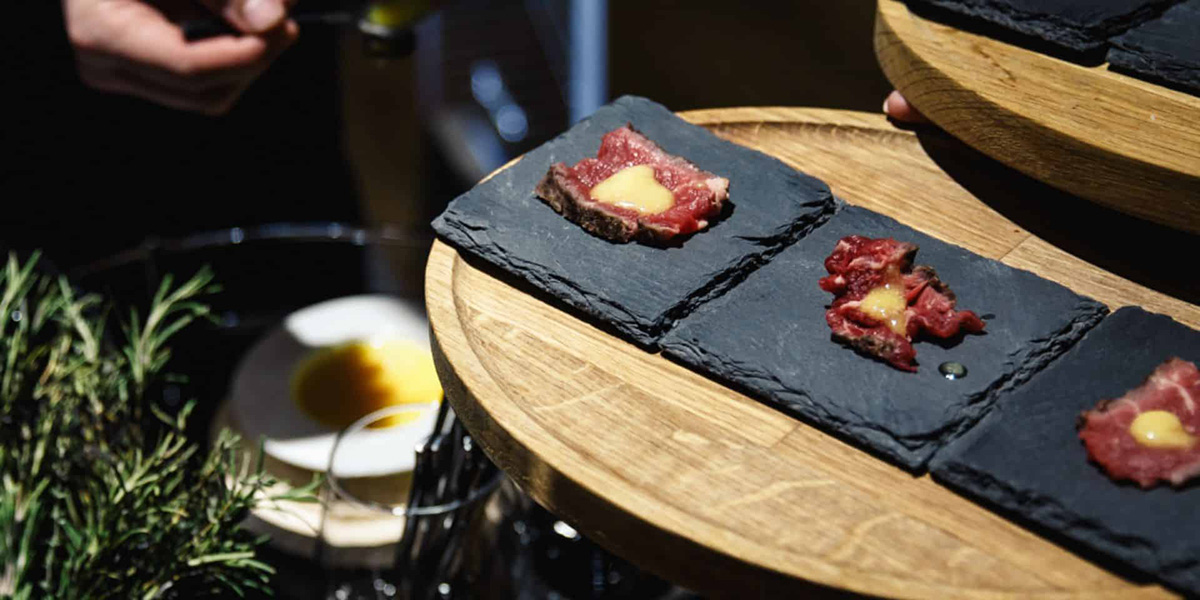 Three small pieces of raw beef topped with sauce served on black slate plates on a wooden platter.
