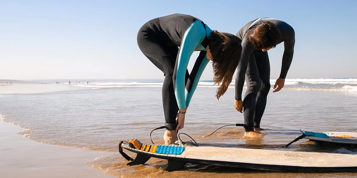 Two people on a beach in wetsuits attaching surfboard leashes to their ankles near the water.