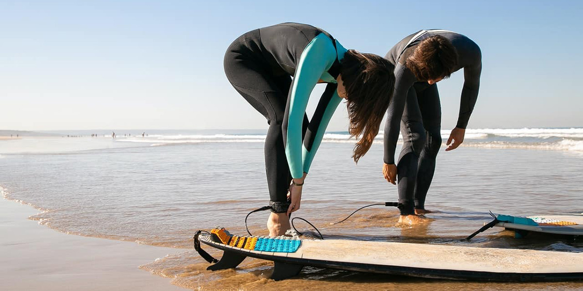 Two people on a beach in wetsuits attaching surfboard leashes to their ankles near the water.