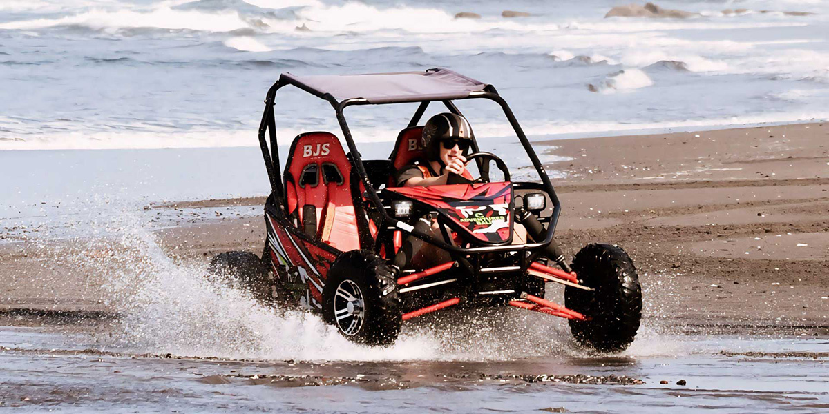 Person driving a red beach buggy splashing through shallow water on a sandy beach.