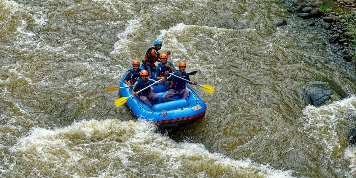Five people wearing helmets and life jackets rafting in a blue inflatable boat on a fast-flowing river.
