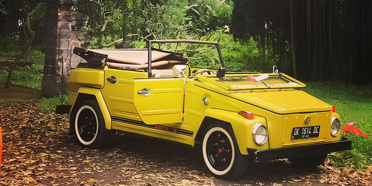 Yellow vintage convertible car with whitewall tires parked on leafy ground near green foliage.