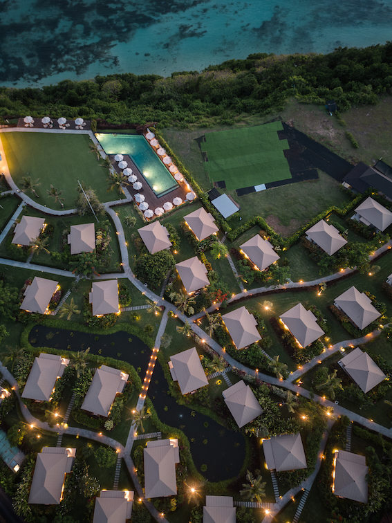 Aerial view of a beachfront resort with illuminated pathways, individual villas, a swimming pool, and lush greenery.