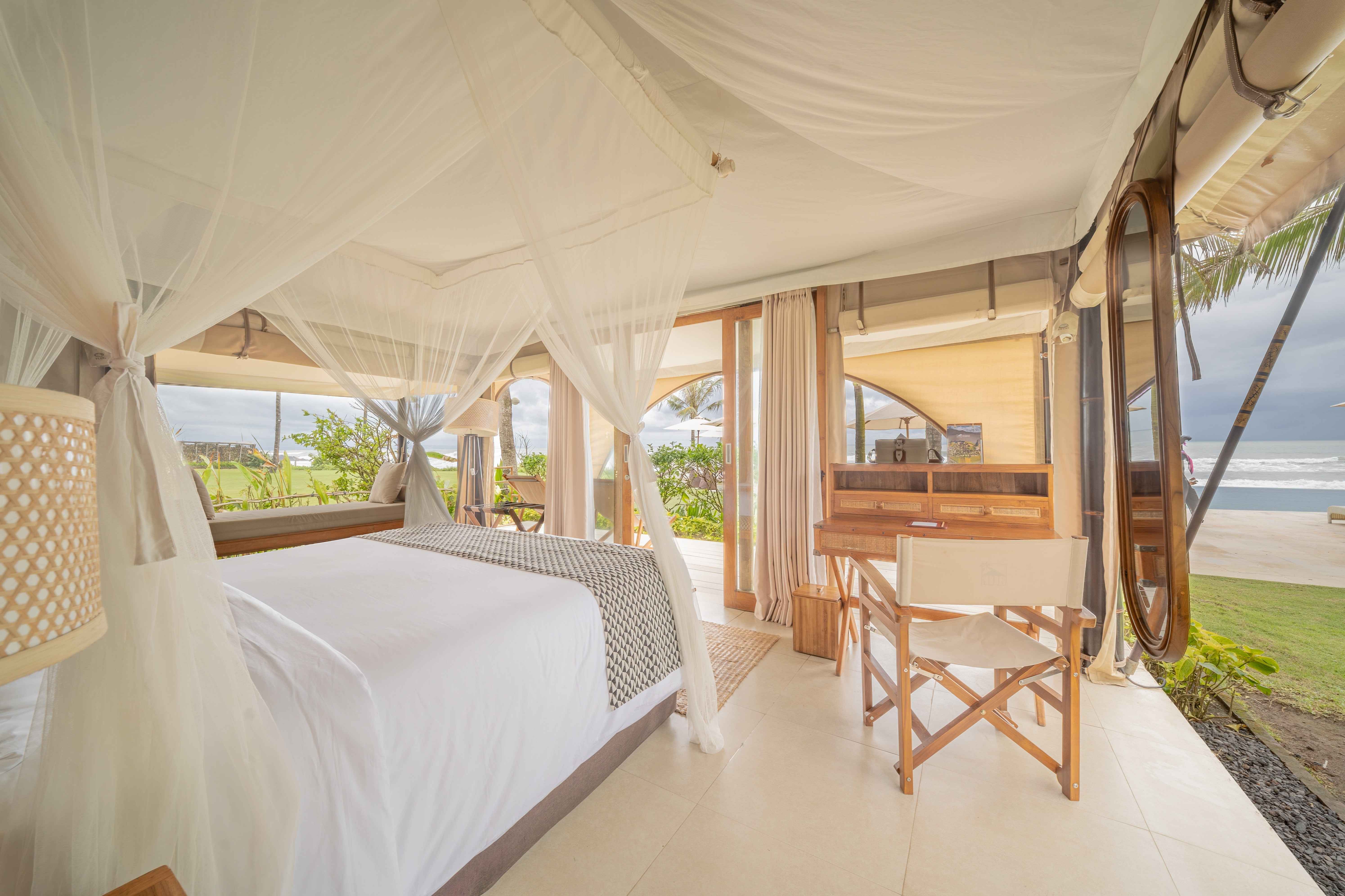 Bright tented bedroom with canopy bed, wooden desk and chair, overlooking greenery and ocean.