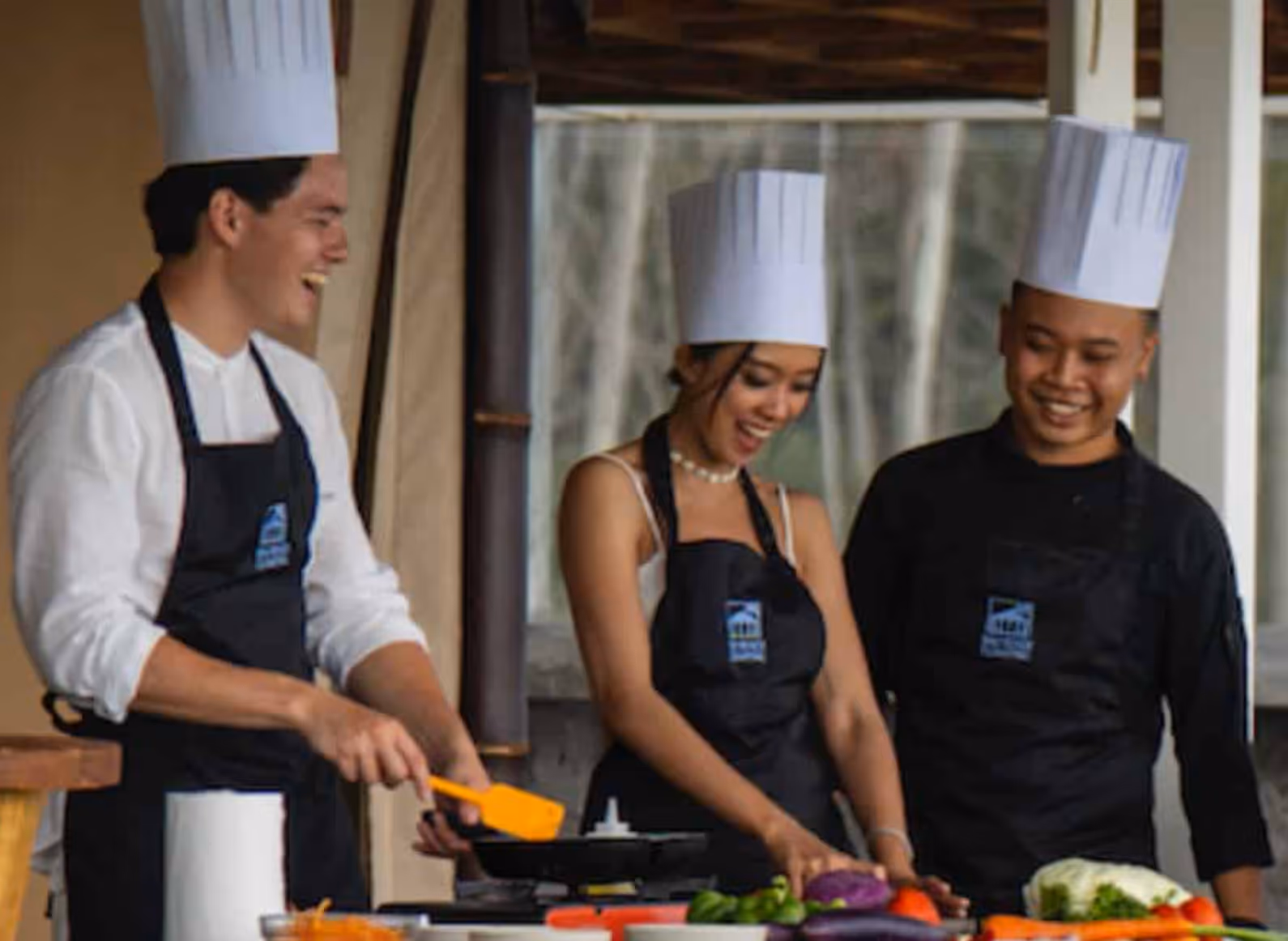 Three chefs in white hats and black aprons smiling and cooking with fresh vegetables on a table.
