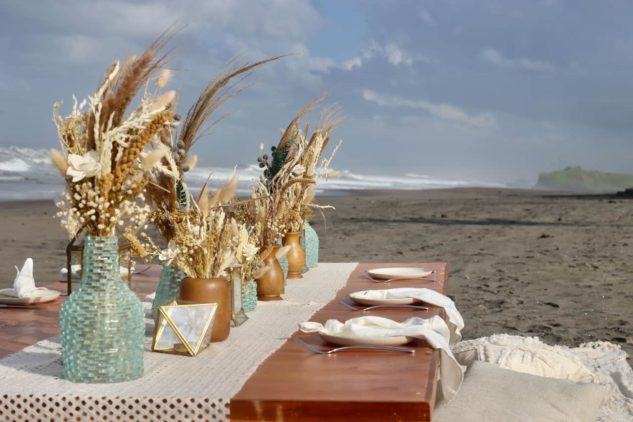 Beachside wooden table set with dried flower arrangements and tableware under a cloudy sky.