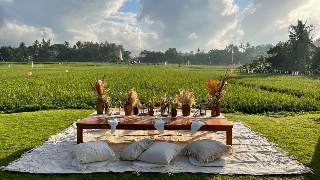 Low wooden table set for picnic with cushions on blanket in grassy field under a cloudy sky.