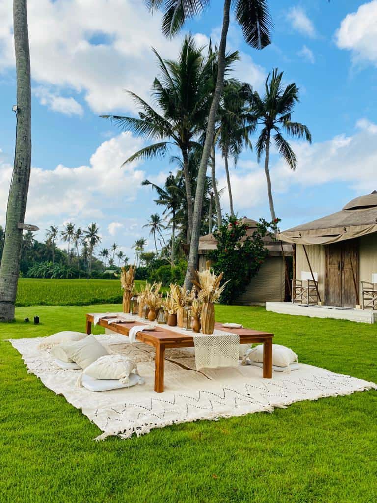 Low wooden picnic table on beige rugs with pillows and dried flower arrangements on green grass under palm trees.