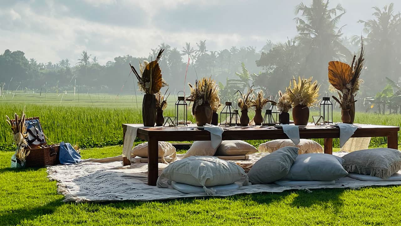 Low wooden table set outdoors on grass with pillows, dried floral arrangements, and lanterns for a picnic.