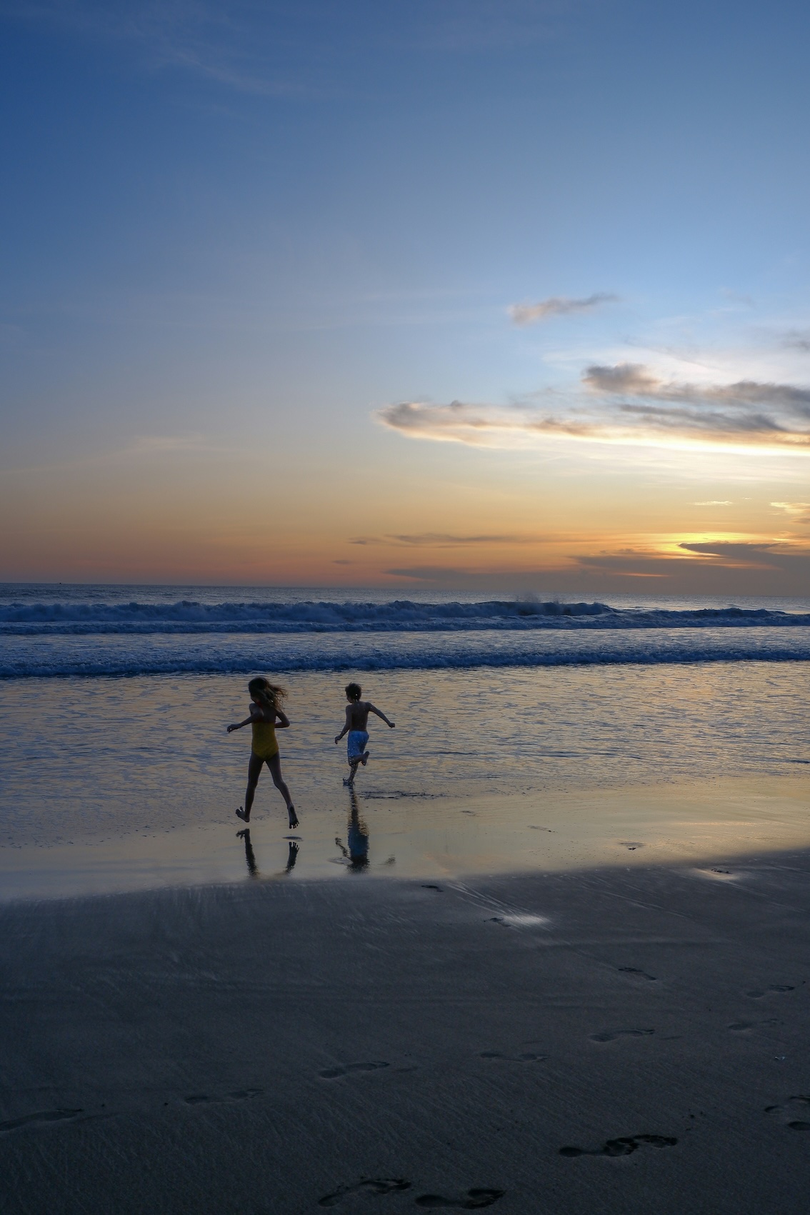 Two children running and playing near the ocean shore at sunset with waves and cloudy sky.