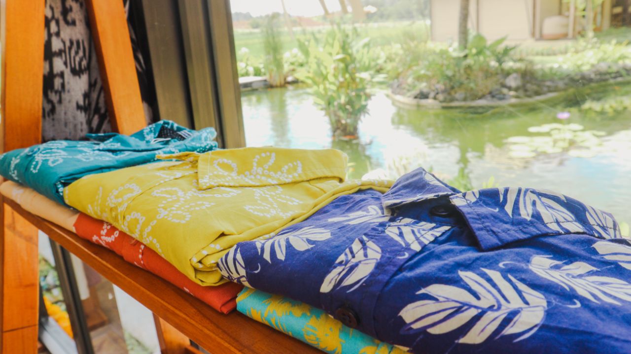 Folded colorful tropical print shirts arranged on a wooden shelf with a pond and greenery in the background.