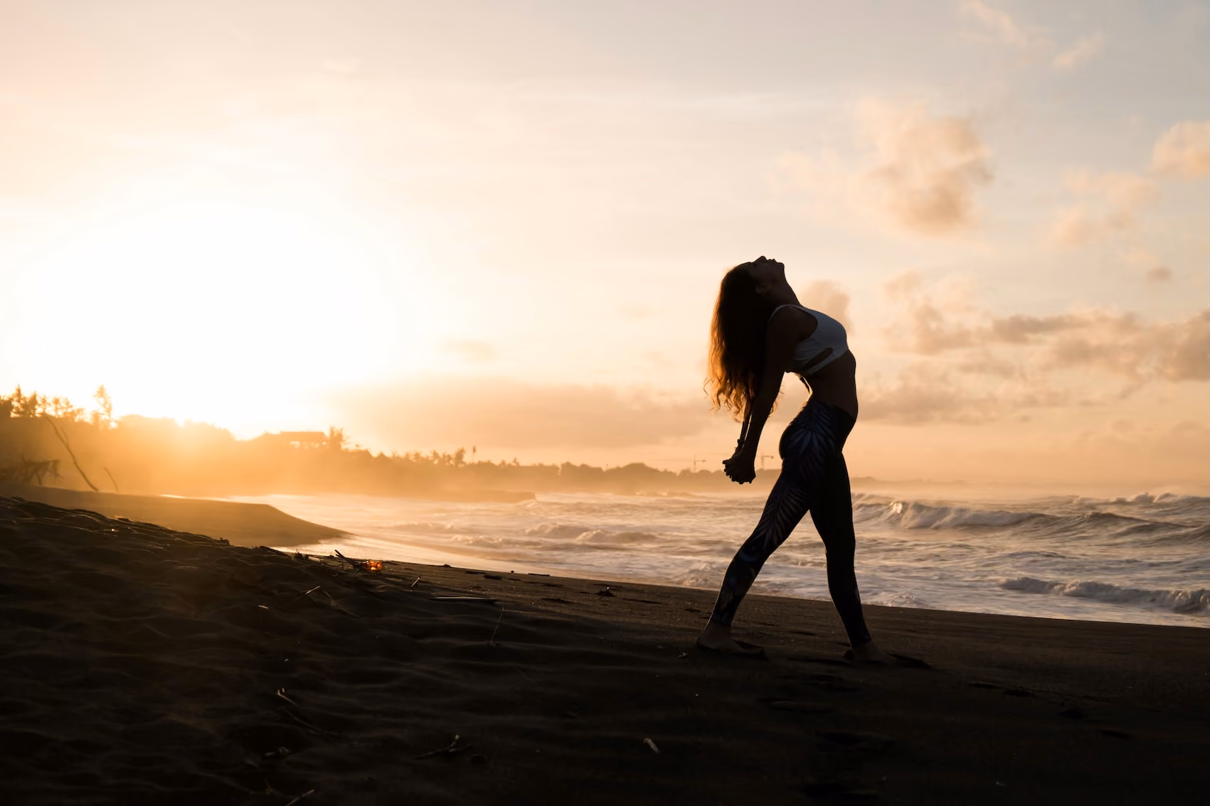 Silhouette of a woman stretching on a beach at sunrise with ocean waves behind her.