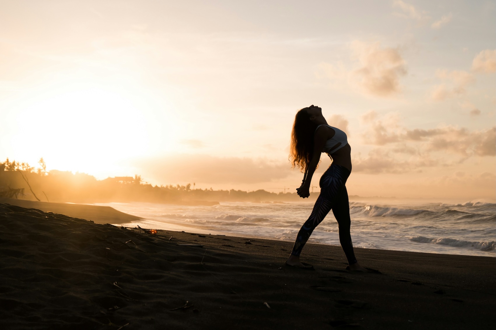 Silhouette of a woman stretching on a beach at sunrise with ocean waves behind her.