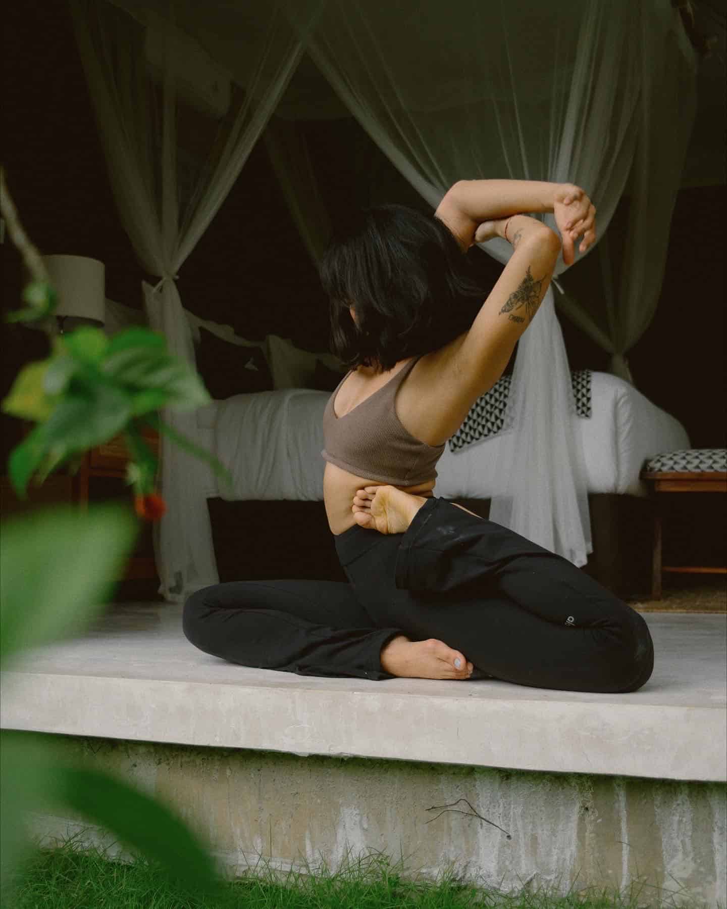 Woman in athletic wear performing a seated yoga pose indoors near a bed with white curtains.