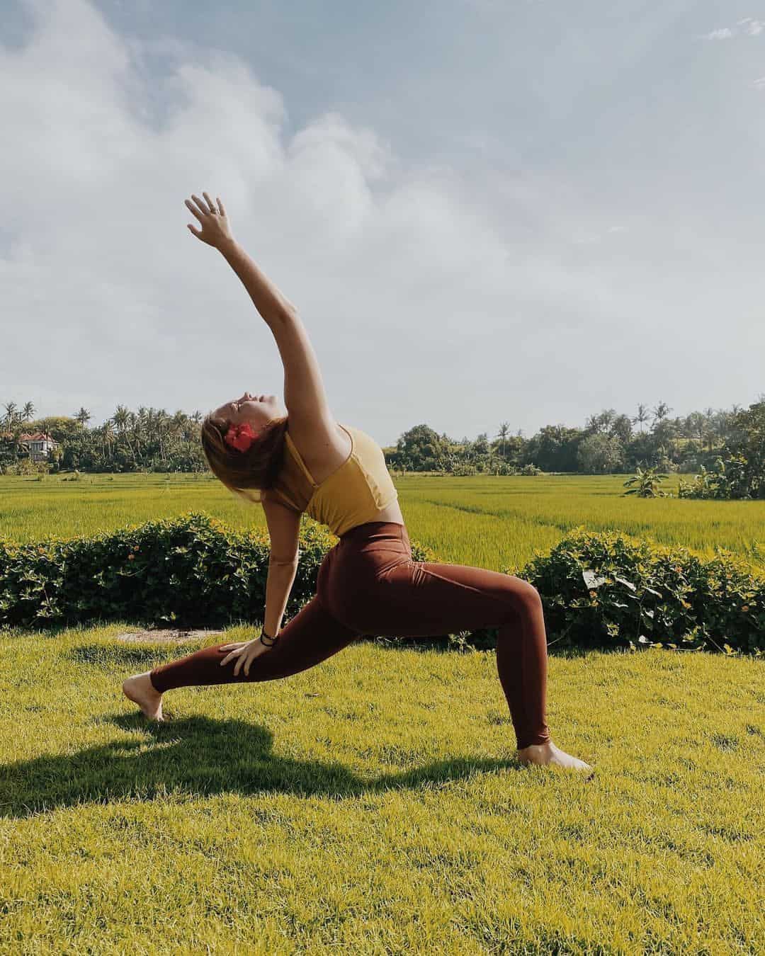 Woman in yellow top and brown leggings doing a yoga stretch on grass with a field and cloudy sky in the background.