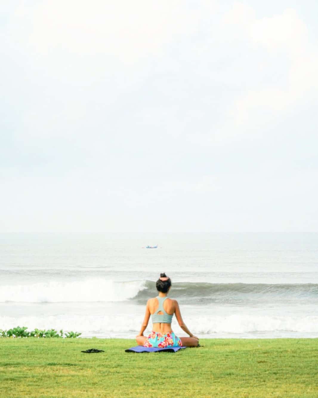 Woman sitting cross-legged on a yoga mat facing the ocean with waves and a distant boat.
