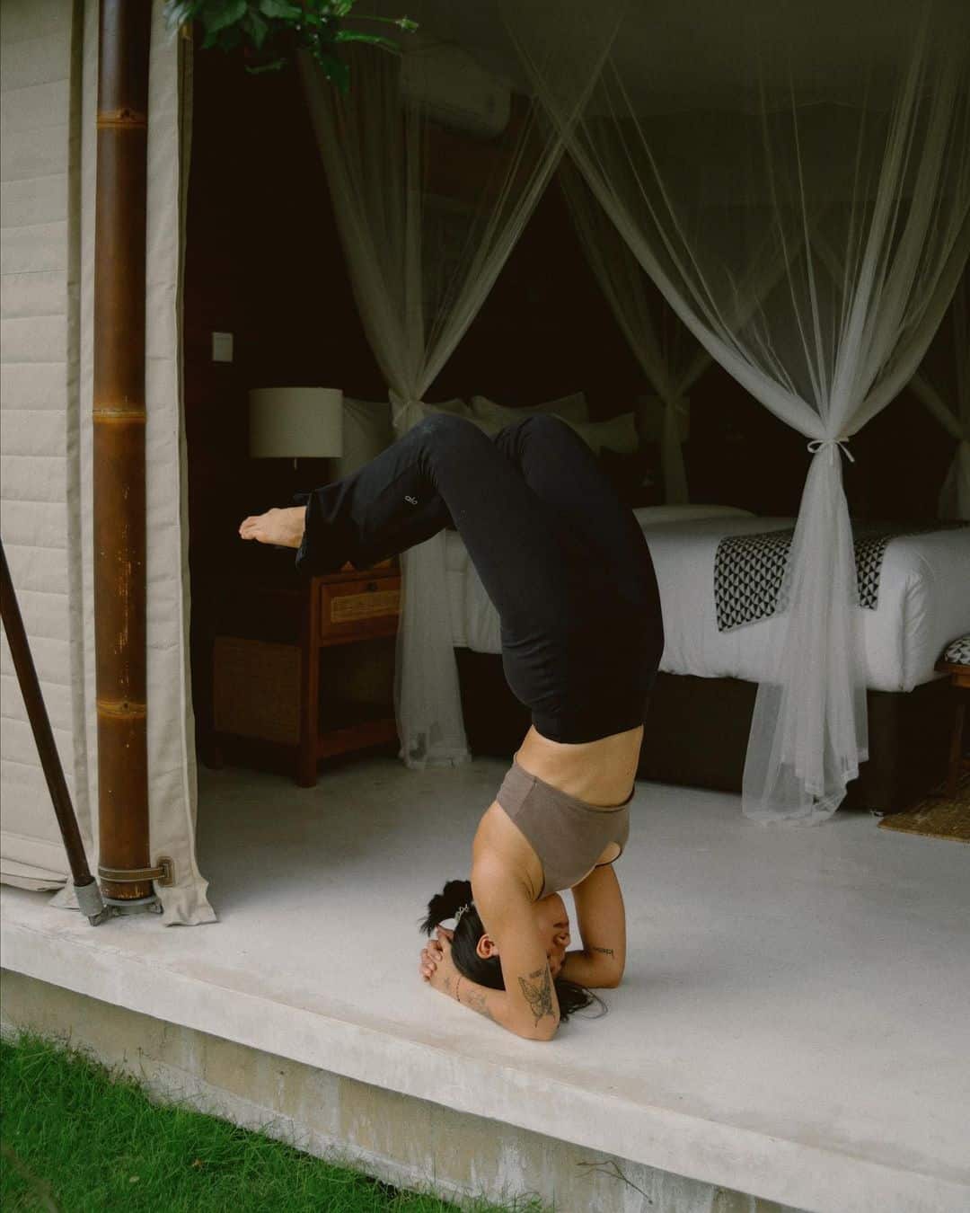 Woman practicing a headstand yoga pose indoors on a light floor near a bed with white curtains.