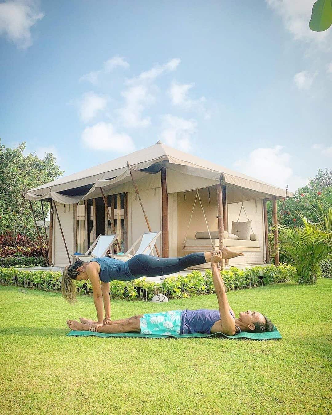 Two people practicing acroyoga on grass in front of a canvas tent with lounge chairs and swing.