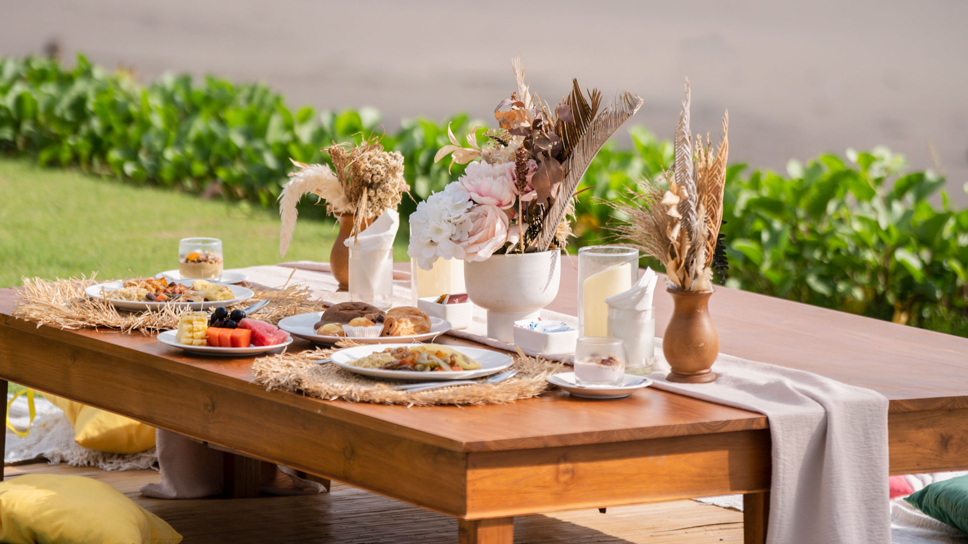 Outdoor wooden picnic table set with breakfast plates, fruit, pastries, and floral centerpieces.