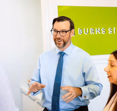 Smiling man in glasses and blue shirt speaking with a woman in an office with a green sign reading 'BURNS SIE' in the background.