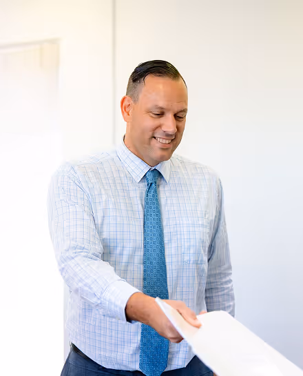 Smiling man in a light checked shirt and blue tie extending his hand forward.