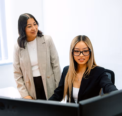 Two professional women collaborating in an office, one seated working on a computer and the other standing beside her.