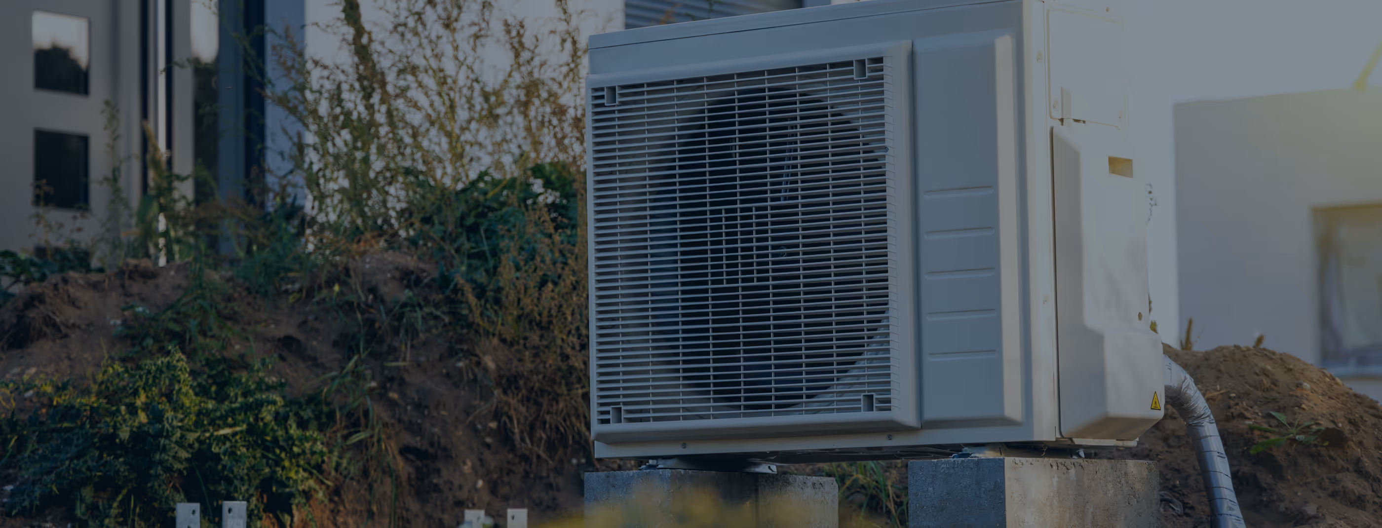Unité de climatisation externe installée près d'un bâtiment avec végétation envahissante alentour.
