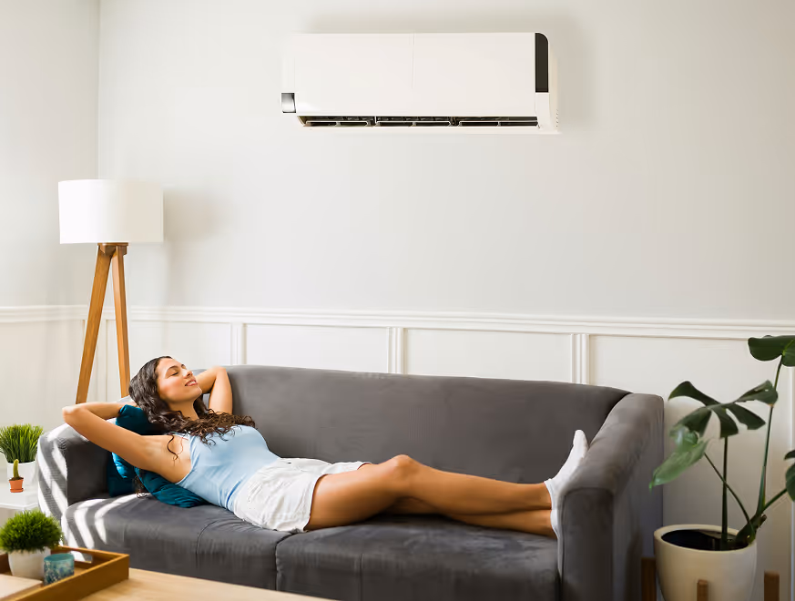 Femme relaxée sur canapé gris, climatiseur mural, plantes vertes, lampadaire bois.