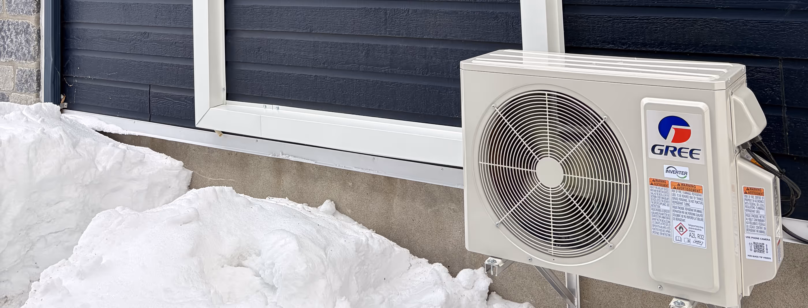 Unité extérieure de climatisation Gree installée près d'une porte de garage noire entourée de neige.