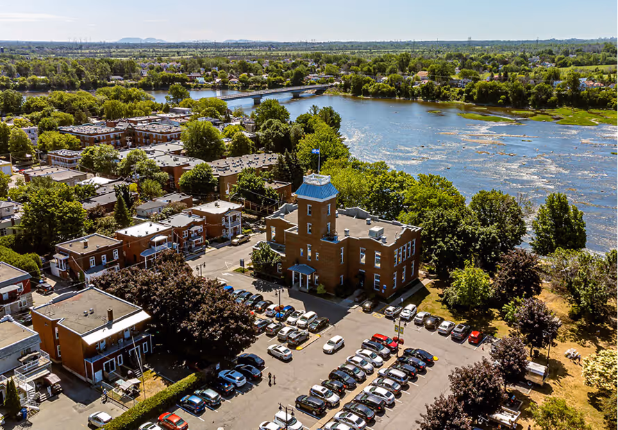 Vue aérienne d'une ville riveraine avec bâtiments historiques, arbres verts et fleuve bleu.