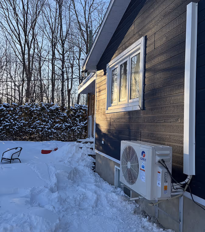 Maison hivernale avec unité de climatisation extérieure entourée de neige et arbres nus.