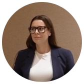 Woman with long brown hair wearing glasses, a white shirt, and a dark blazer against a beige background.