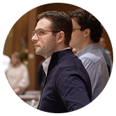Man with glasses and a beard wearing a navy jacket, sitting thoughtfully in a meeting or classroom setting.