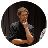 Man with curly hair and beard wearing a black shirt, resting chin on hand in thoughtful pose against a dark background.