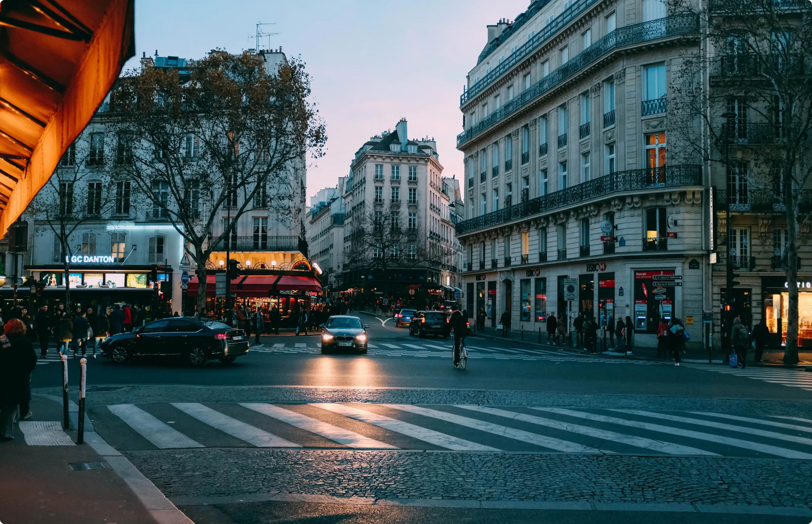 Evening city street scene with pedestrians, cars, a cyclist, and historic buildings under a dusky sky.