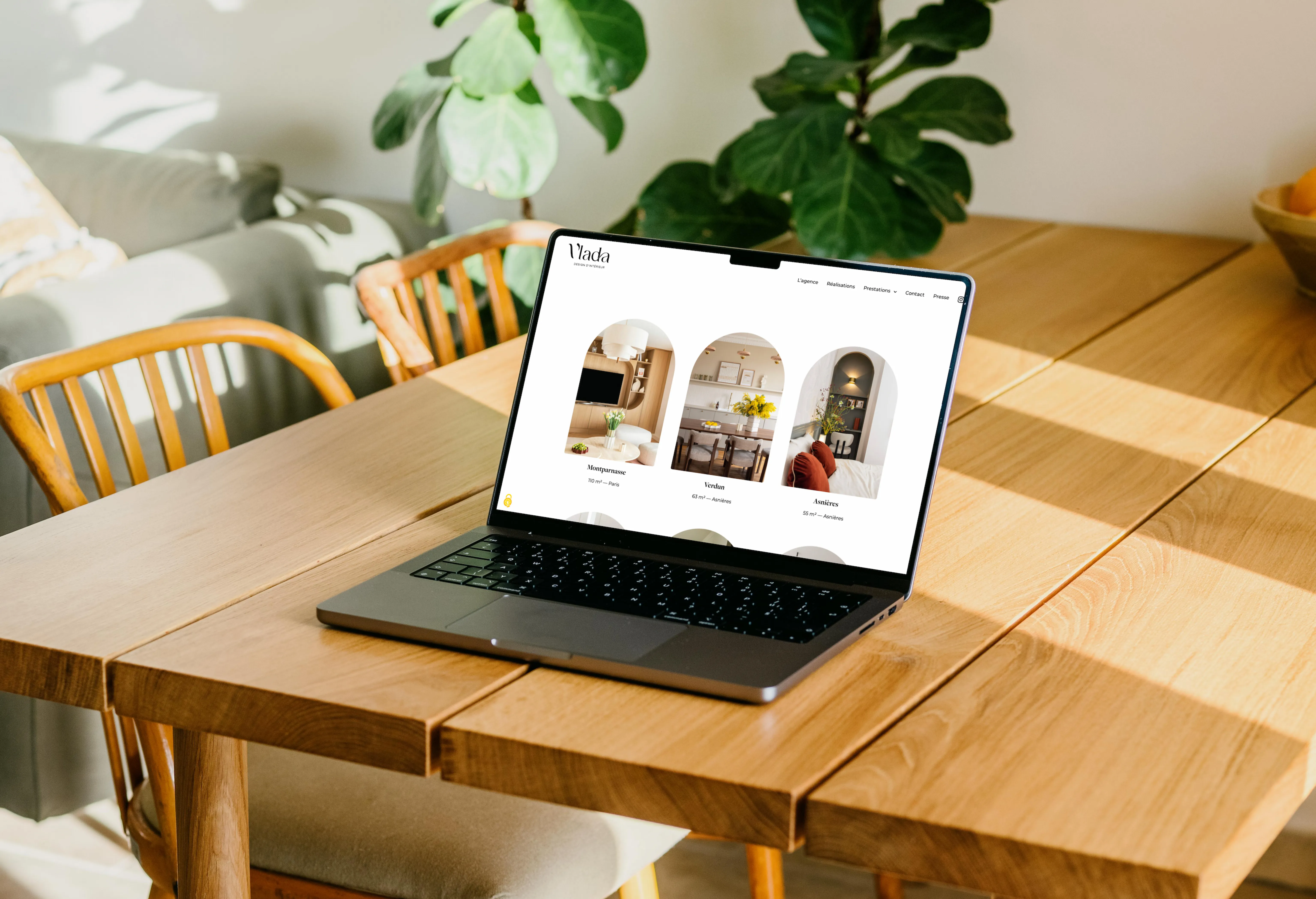Open laptop displaying interior design website on wooden table with chairs and green plant in background.