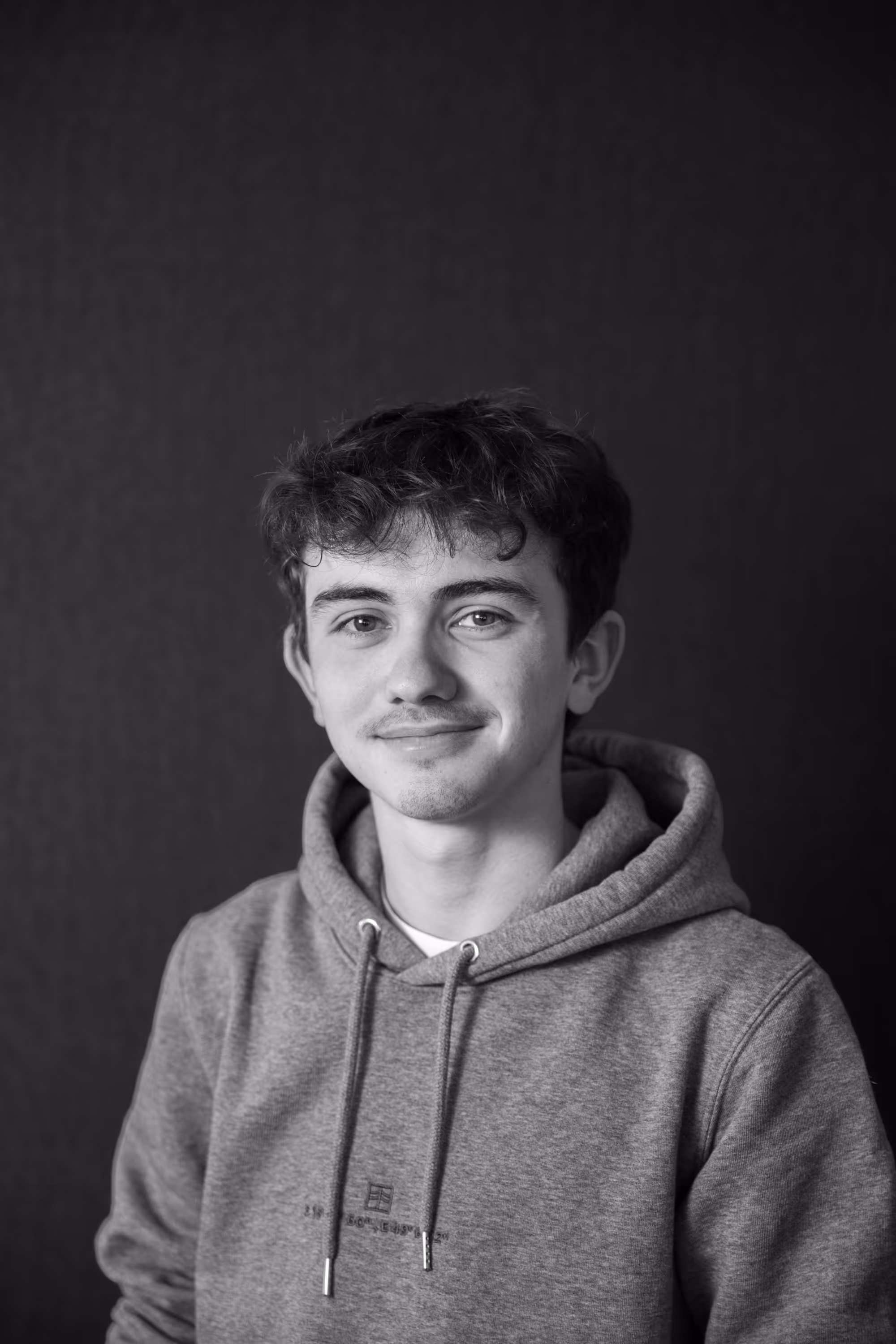 Young man with curly hair wearing a hoodie smiling gently against a dark background.