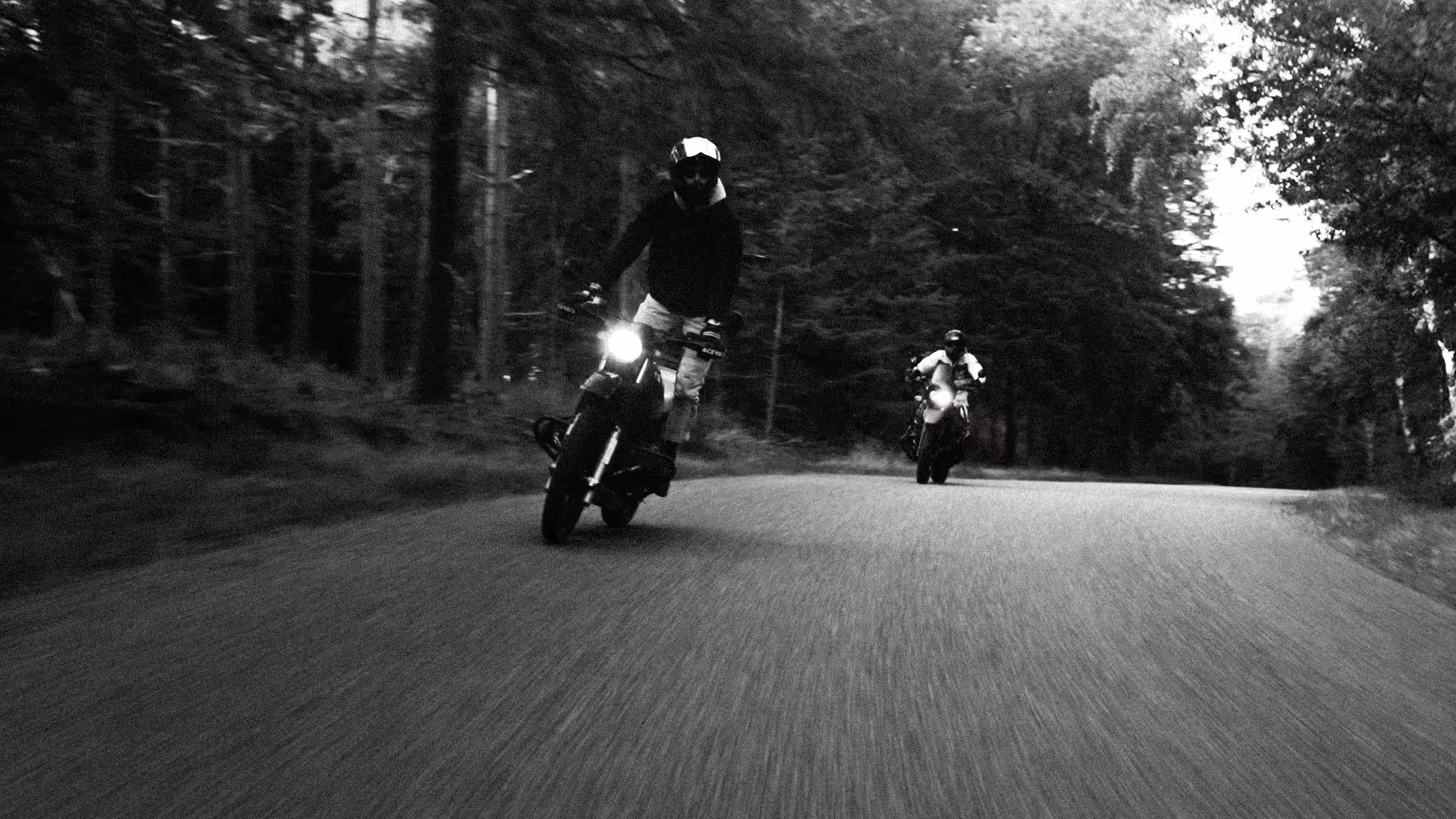 Two motorcyclists riding on a forested road, with the closer rider performing a wheelie.