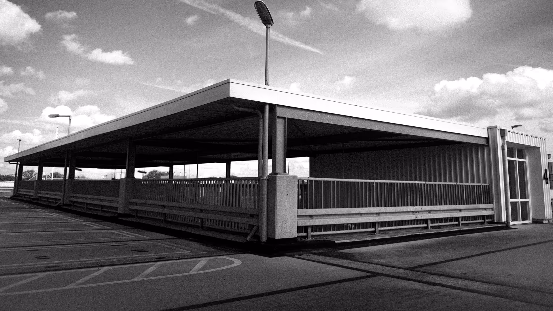 Black and white photo of an empty rooftop parking structure with a covered area and street lamps under a partly cloudy sky.