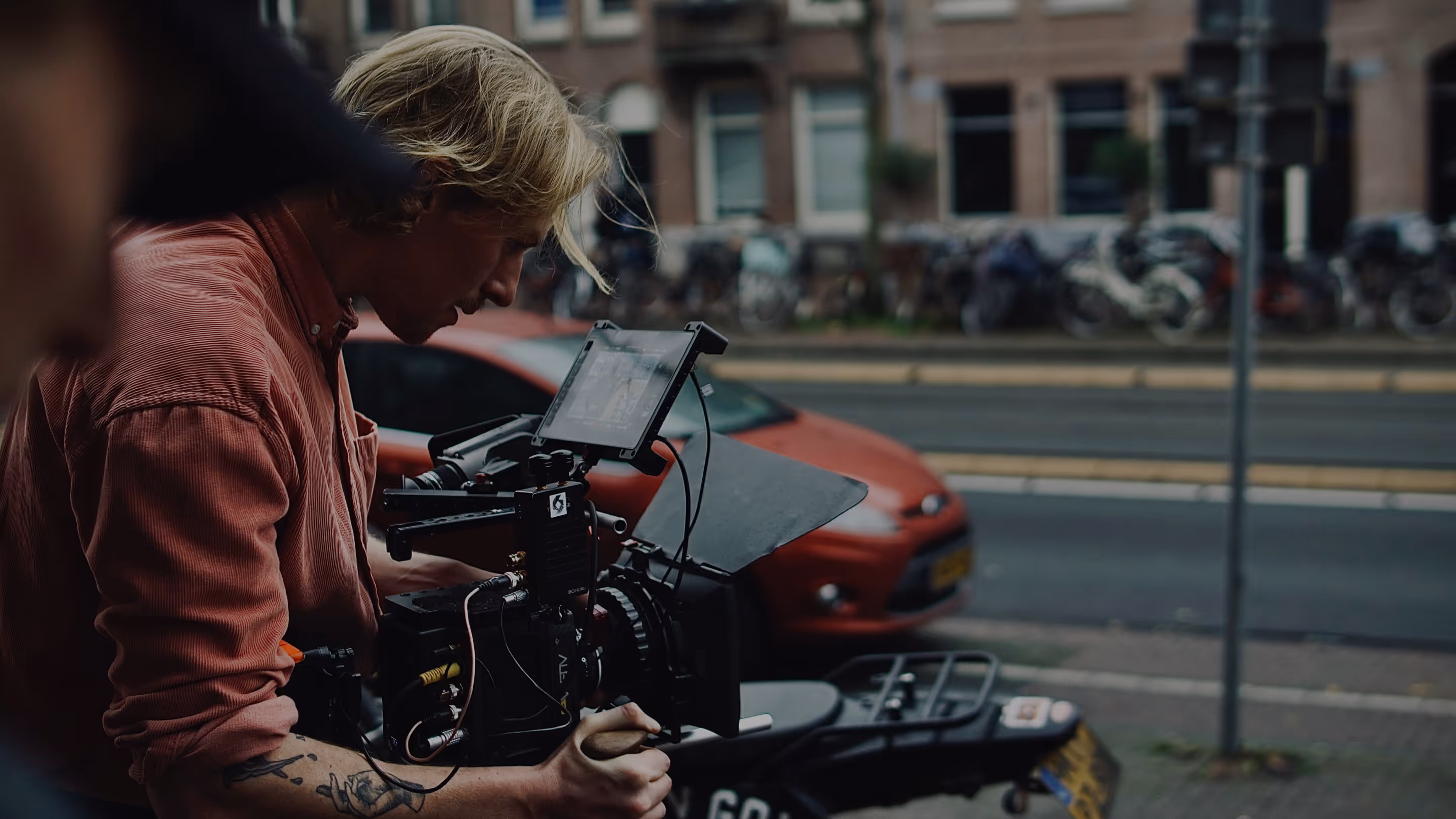 Cameraman with blond hair and tattoo operating a professional video camera on a street.