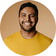 Smiling man with short dark hair and beard wearing a yellow shirt against a brown circular background.