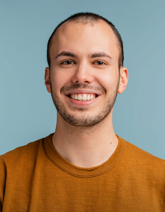 Smiling young man with short hair and beard wearing a brown shirt against a blue background.