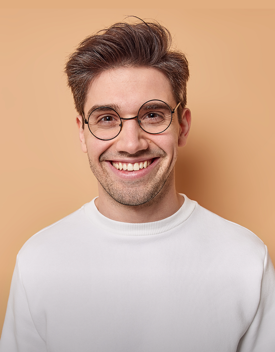 Smiling young man with round glasses and styled brown hair wearing a white sweater against a beige background.