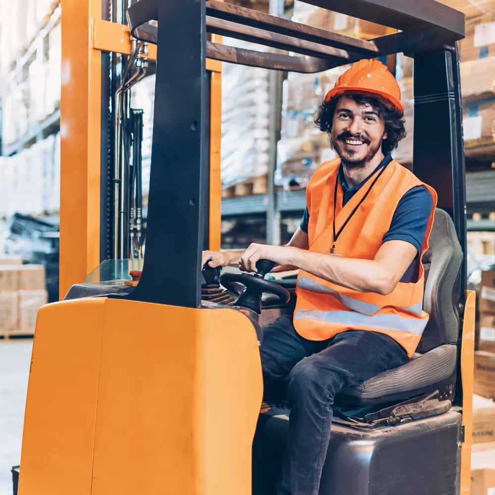 Mann mit orangefarbener Schutzweste und Helm lächelt beim Bedienen eines Gabelstaplers im Lager.