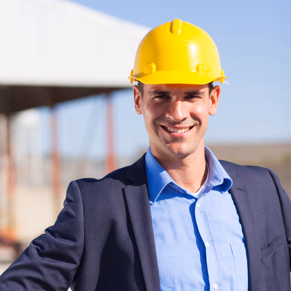 Mann mit blauem Hemd, Anzug und gelbem Helm lächelt auf einer Baustelle.